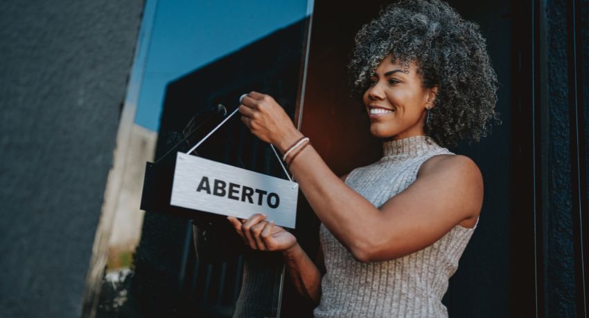 Mulher sorridente pendurando placa de “Aberto” na porta da loja, marcando o início das operações.